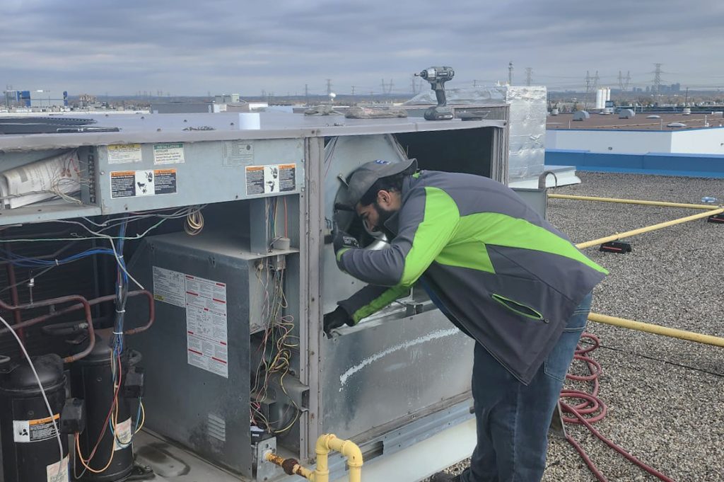 An HVAC specialist in a green and gray jacket performing maintenance on a rooftop unit against an urban backdrop.